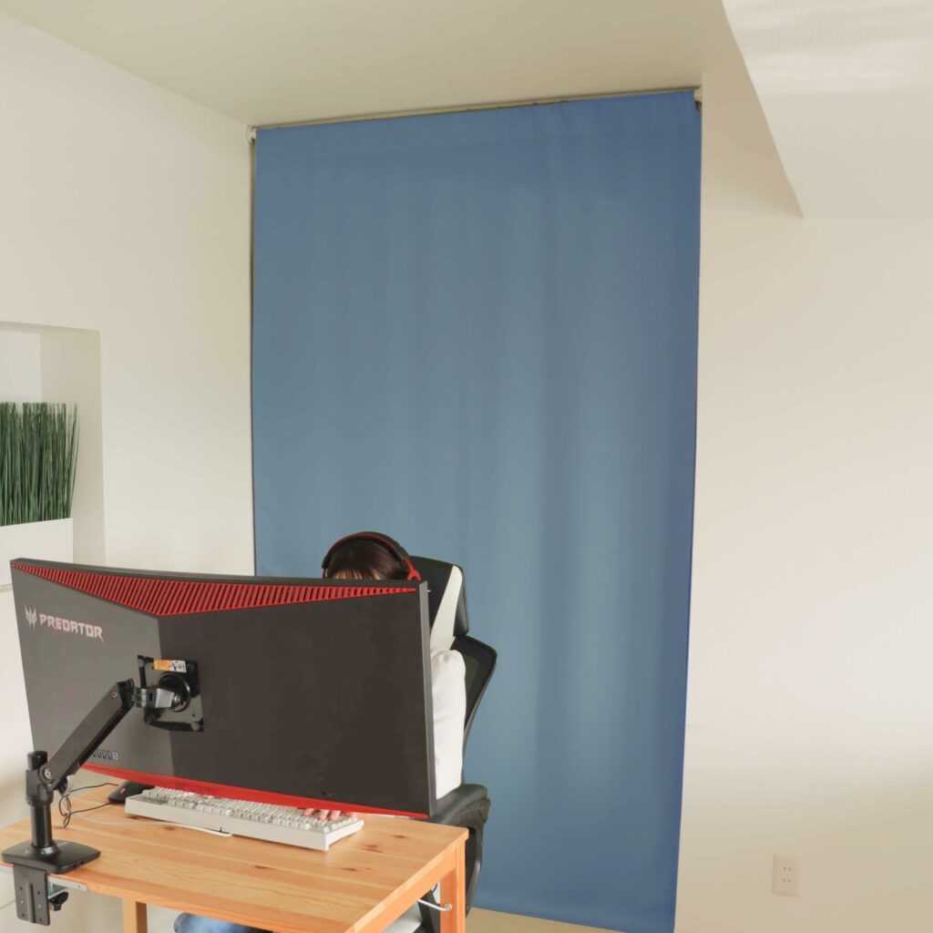 間仕切りとして突っ張り棒で設置できる防音カーテンパーテンウィンターブルーがゲームをしている女性の背面の窓に音漏れの防音対策として設置されている